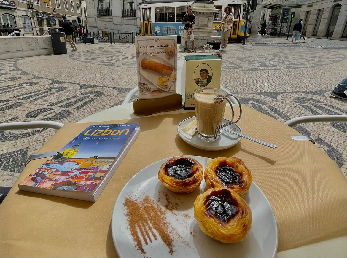 Tre pastéis de nata drysset med kanel og en mælkeagtig kaffe på et cafebord, med en sporvogn på det mønstrede fortov bagved